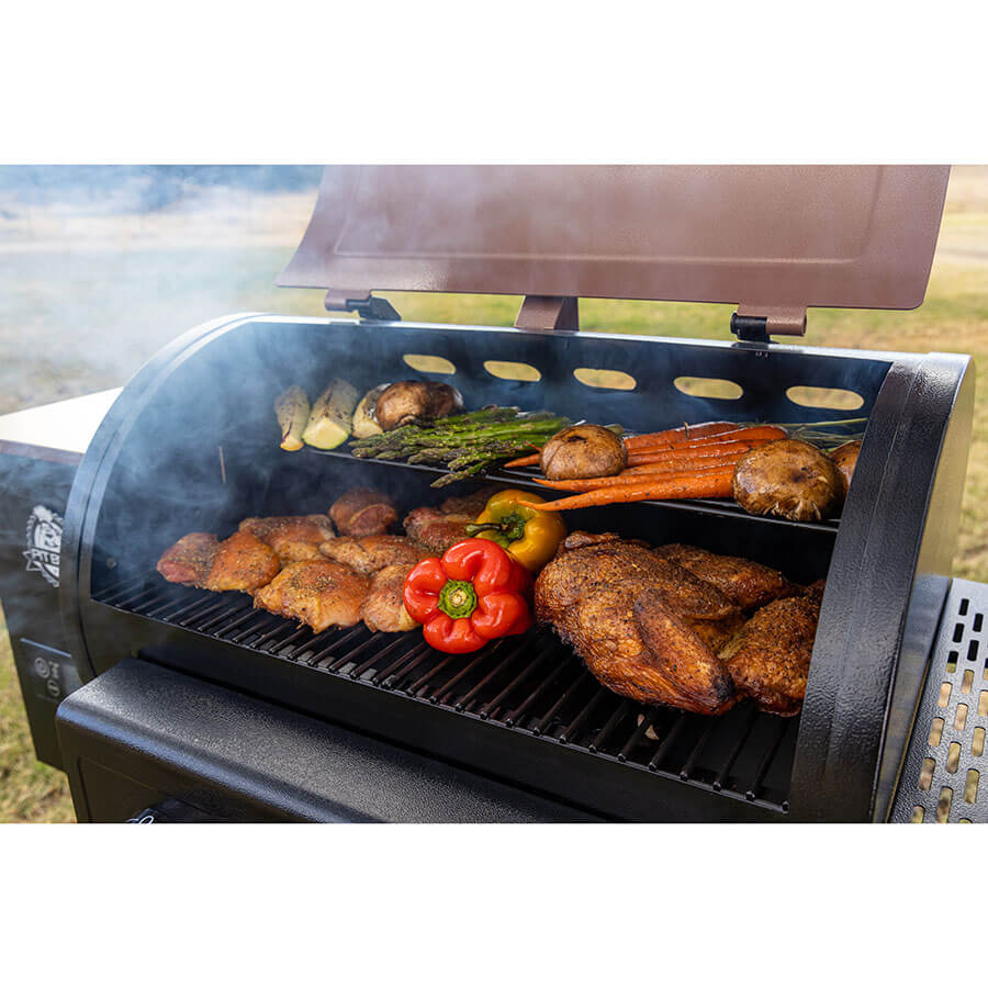 Close up of meats and veggies cooking on both grilling racks with smoke coming out. Grill outside in a yard.