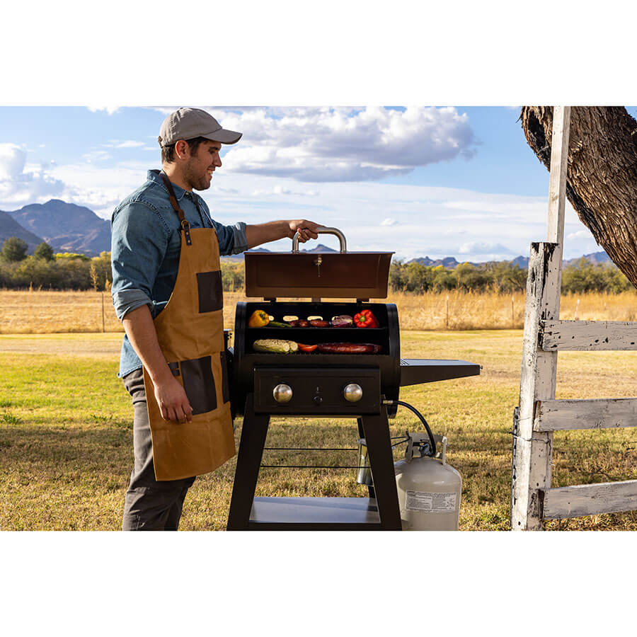 Man holding grill hood open. Grill outside with mountains and field behind. Meats and veggies cooking.