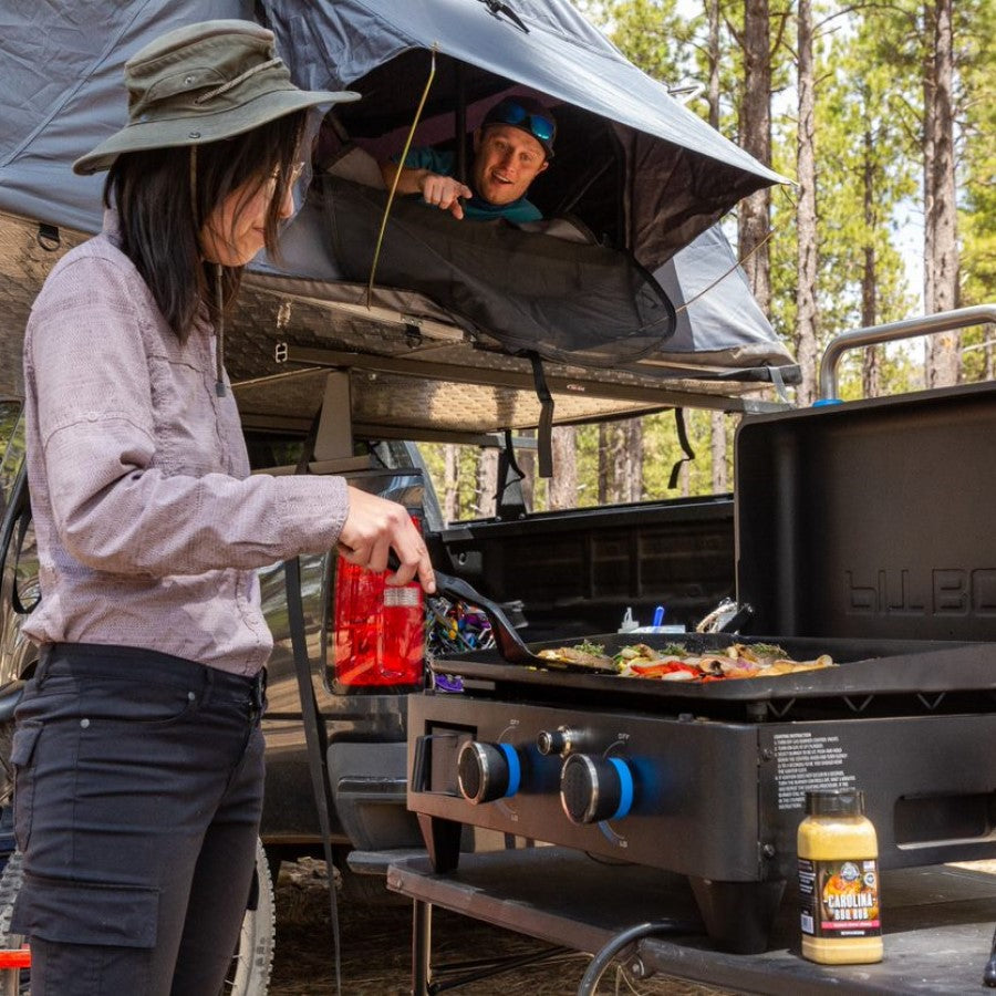 lifestyle_4, Black griddle with blue and silver accents. Large blue "Pit Boss" logo across front.  Picture shows woodsy camping scene