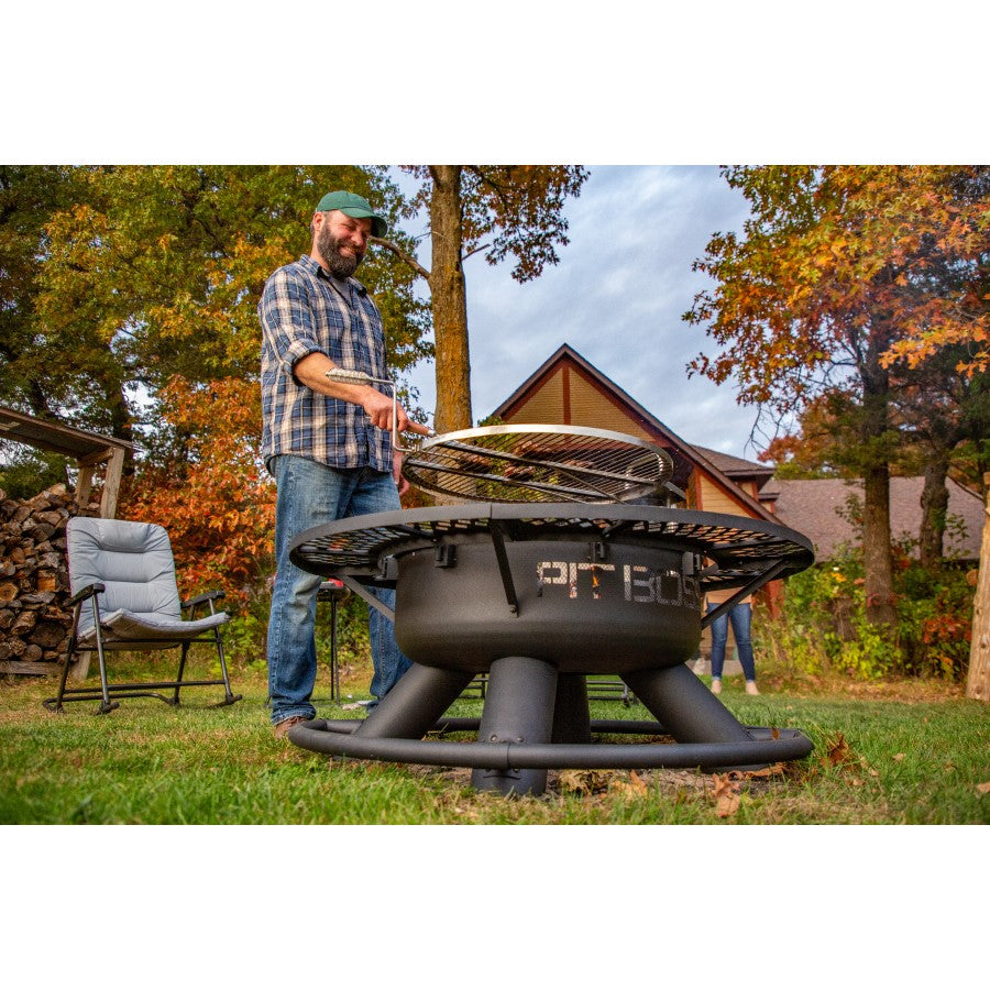 lifestyle_6, Black fire pit with round shape and black lettering: Pit Boss. Man standing over fire pit cooking meat